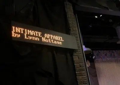 A black LED captioning screen hanging next to a stage. Orange text on the screen reads "Intimate Apparel by Lynn Nottage."